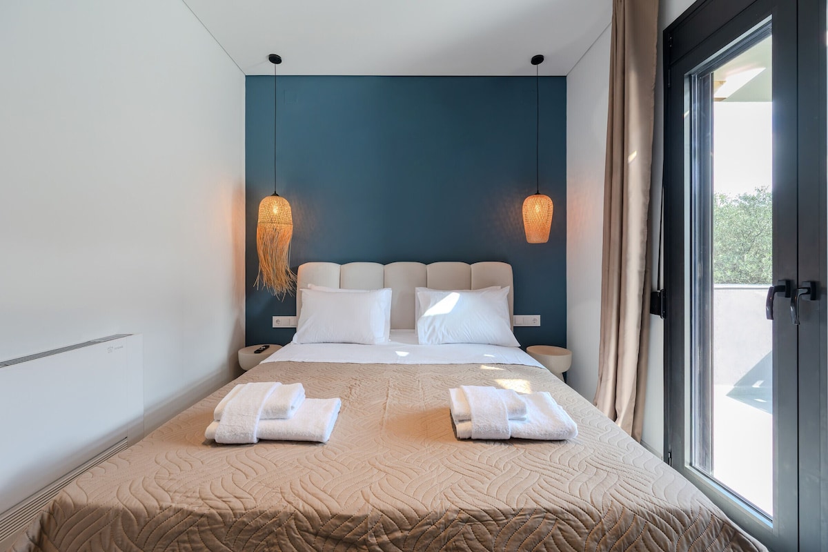 A modern hotel room with a large bed, two bedside tables, a window with curtains, and a view of the outdoors.