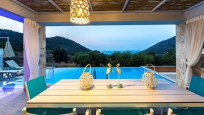An outdoor dining area with a view of the sea and mountains, a pool, and lounge chairs. - Sugar Premium Villa (Mylopotamos)