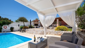 A poolside area with a glass table, wicker furniture, and a stone wall. - Villa Plumeria Flower (Chania)