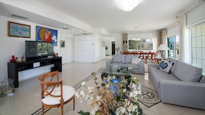 A spacious living room with a sofa, a glass coffee table, and a television on a black stand. There is a dining area with a table and chairs in the background. - Villa Plumeria Flower (Chania)