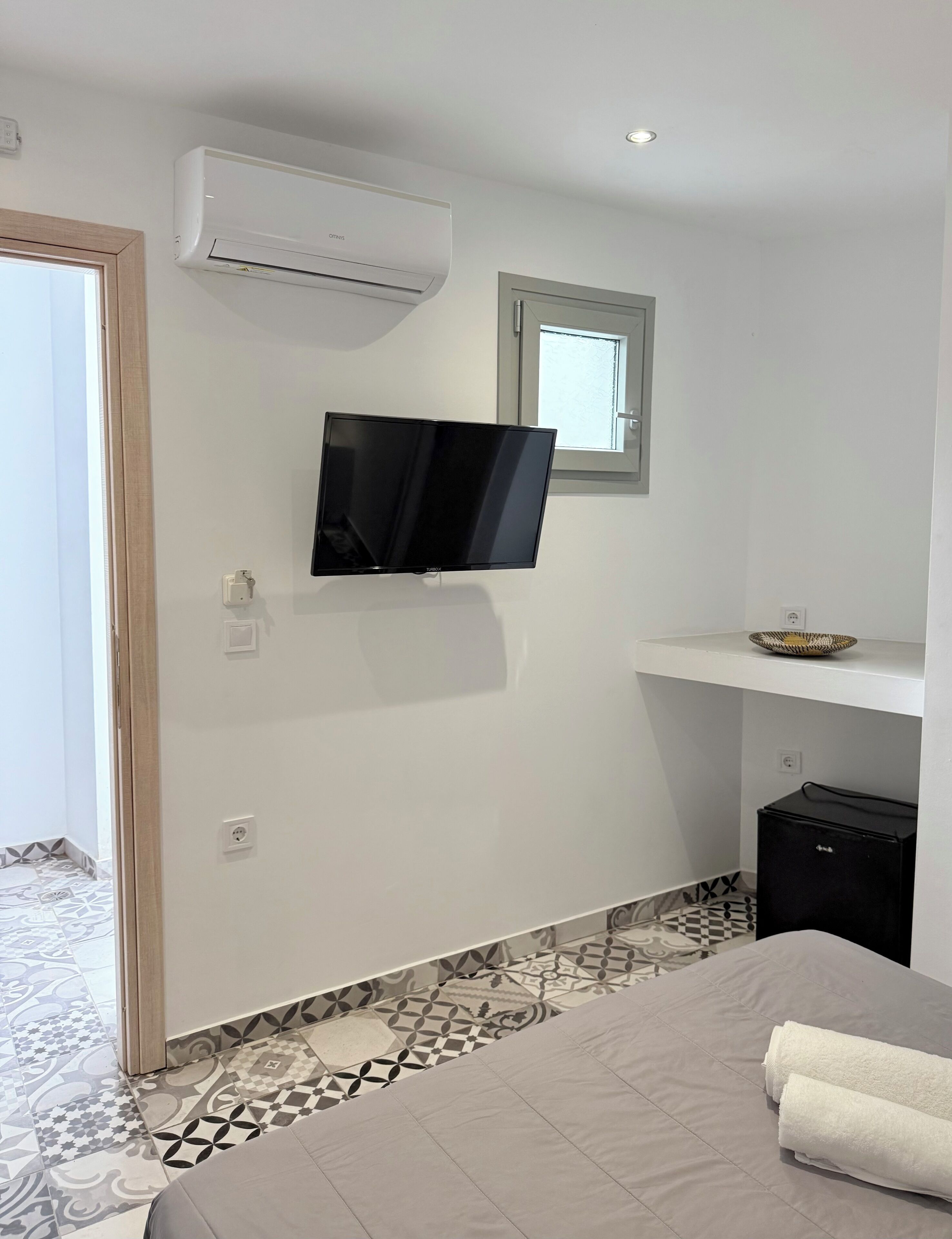 Un dormitorio con cama, televisor montado en la pared, aire acondicionado, ventana pequeña y piso de baldosas.