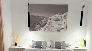 A neatly made bed with two rolled towels, a large framed picture of a coastal town, and a bedside table with a vase of dried flowers. - Fira Basement Studios (Santorini)