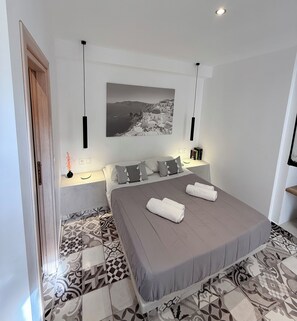 A modern bedroom with a large bed, patterned floor tiles, a wall-mounted picture, bedside lamps, and a door. - Fira Basement Studios (Santorini)