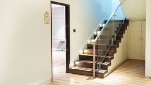 Una escalera moderna con barandillas de vidrio y escalones de madera que conduce a un espacio con suelo de madera y paredes blancas.
