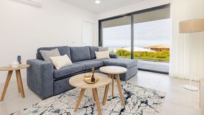 Ett modernt vardagsrum med en grå soffa, två runda träbord och ett stort fönster med utsikt över grönska och byggnader.