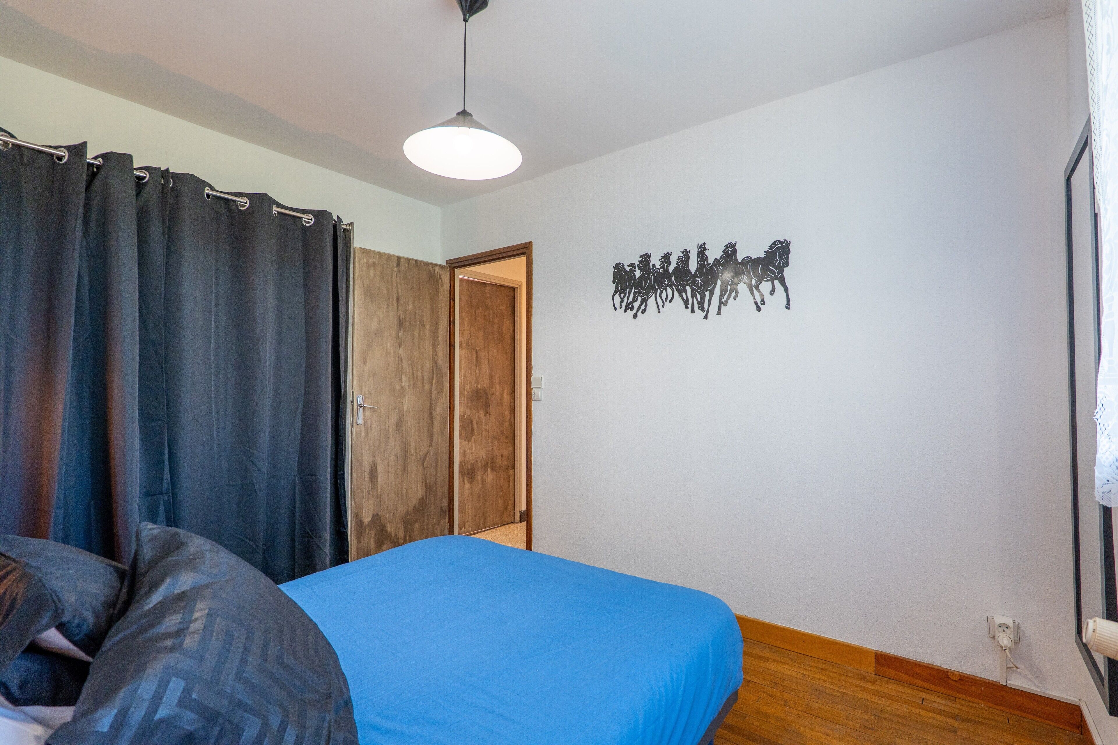 A bedroom with a bed, curtains, a wooden wardrobe, a wall-mounted artwork of horses, and a ceiling light.