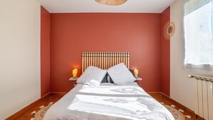 Een slaapkamer met een bed, houten hoofdeinde, nachtlampjes en een raam met kantgordijnen.