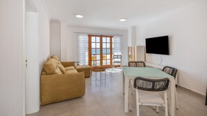 A living room with a sofa, a dining table with chairs, a TV mounted on the wall, and a view through a window. - Apartment 'Playa Delphin 403' with Sea View, Balcony and Wi-Fi (Puerto de Naos)