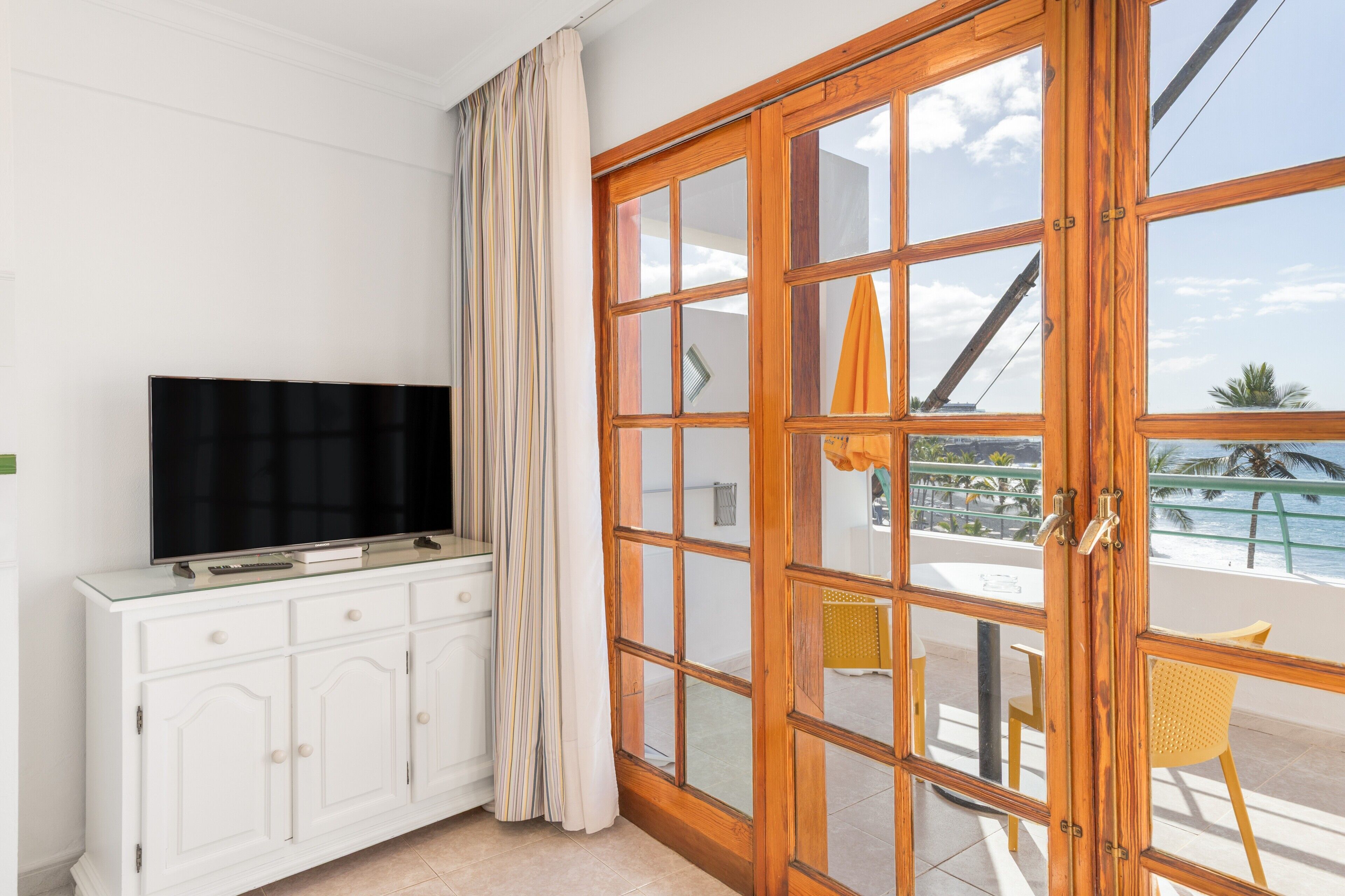 A room with a television on a white cabinet, a sliding glass door to a balcony with a chair and a yellow umbrella, and a view of the ocean.