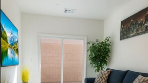A living room with a dark blue sofa, a patterned rug, a sliding glass door leading to a brick patio, and a cityscape painting on the wall. - BRAND NEW Comfy Guest House in a Great Location (Henderson)