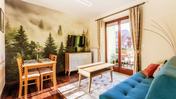 Un salon avec un canapé bleu, une table à manger en bois et des chaises. On y trouve un téléviseur posé sur un meuble, et une vue sur l’extérieur grâce à une porte vitrée.