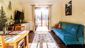 Un salon avec un canapé bleu, une table à manger avec des chaises, un téléviseur posé sur un meuble, et une vue sur un espace extérieur, visible à travers une porte vitrée.
