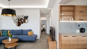 Una sala de estar moderna con un sofá azul, una mesa de centro redonda de madera y una lámpara colgante negra. Hay una zona de cocina con armarios de madera y un televisor empotrado.