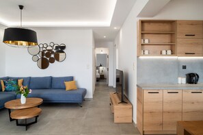A modern living room with a blue sofa, a round wooden coffee table, and a black pendant light. There is a kitchen area with wooden cabinets and a built-in TV. - Zen Apartments (Rethymno)