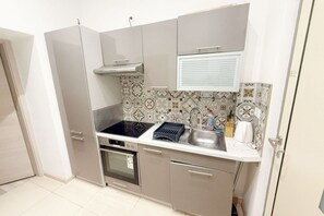 Une cuisine de taille réduite, équipée d’une cuisinière, d’un four, d’un évier et d’une fenêtre avec des stores.