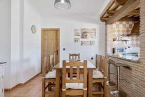 A dining area with a wooden table and chairs, a kitchen with a brick wall, and a hanging light fixture. - Holiday Home 'Villa' with Private Pool, Wi-Fi and Air Conditioning (Conil de la Frontera)