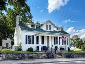 Une maison blanche à deux étages, avec un toit vert, un porche à l’avant et un drapeau américain.