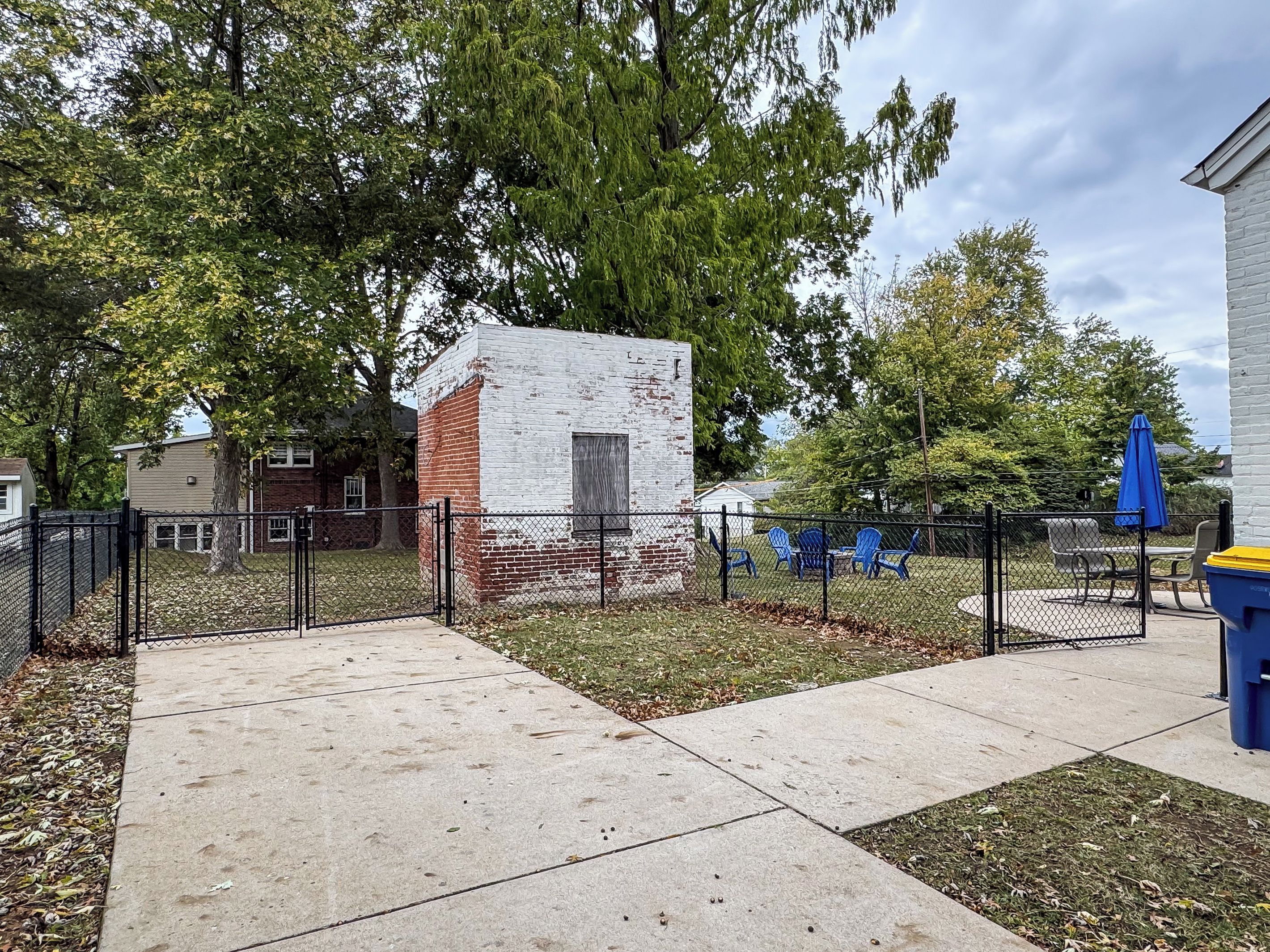 A fenced outdoor area with a small brick structure, a concrete pathway, and blue chairs.