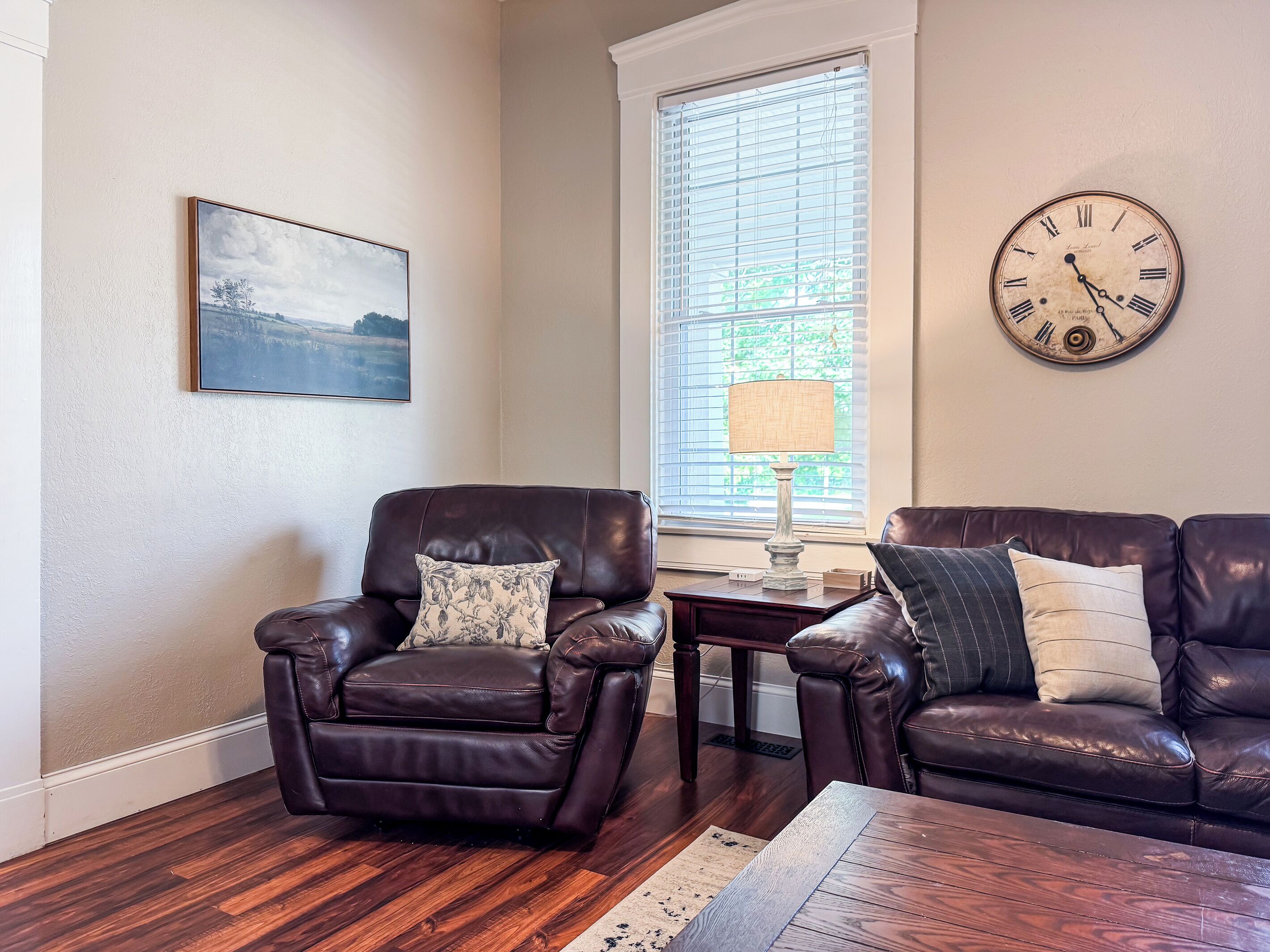 A living room with two leather sofas, a wooden coffee table, a side table with a lamp, a clock on the wall, and a framed landscape painting.