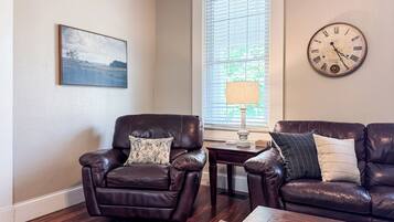 A living room with two leather sofas, a wooden coffee table, a side table with a lamp, a clock on the wall, and a framed landscape painting.