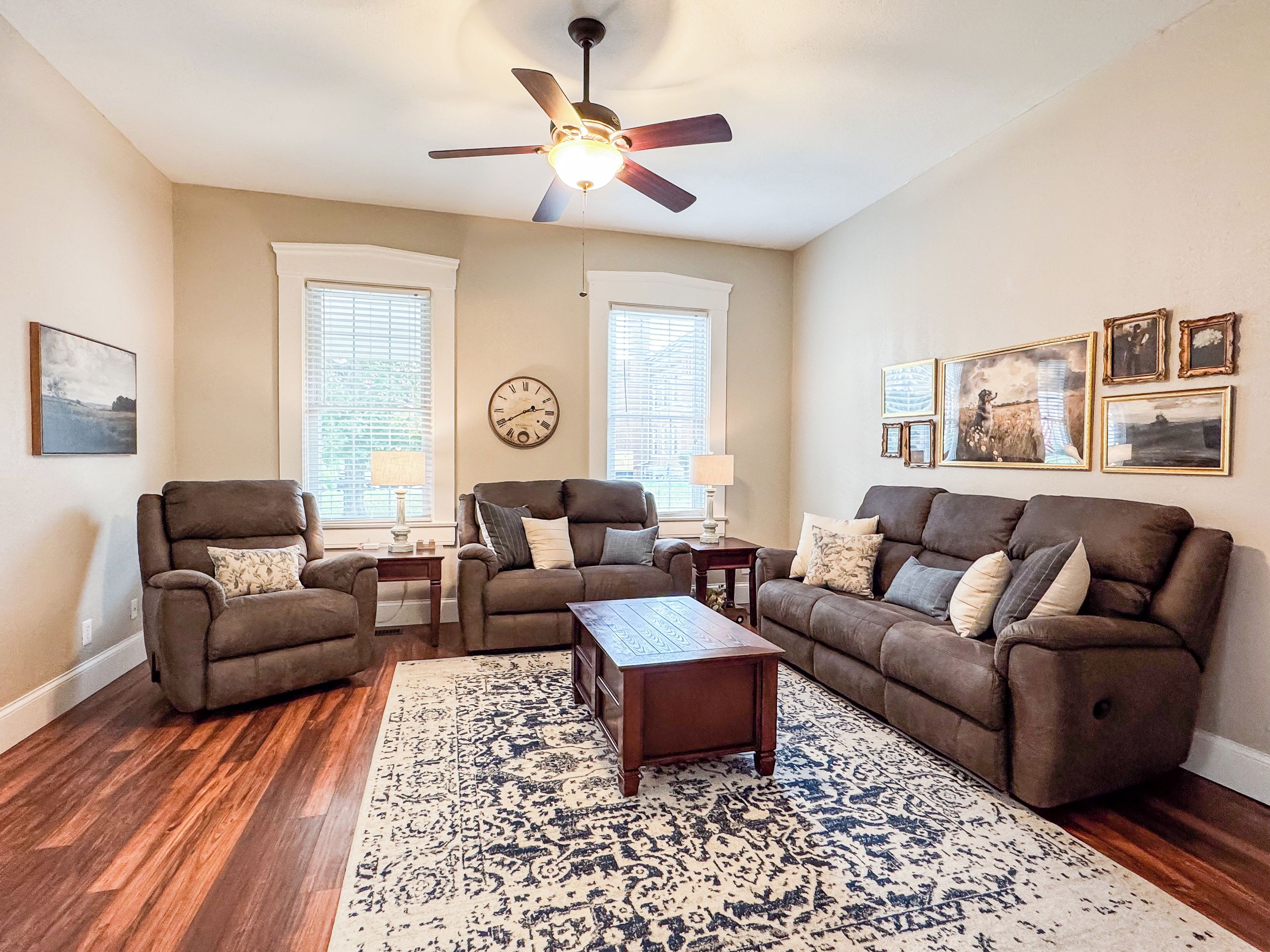 A living room with a ceiling fan, two windows with blinds, a clock, and a rug.