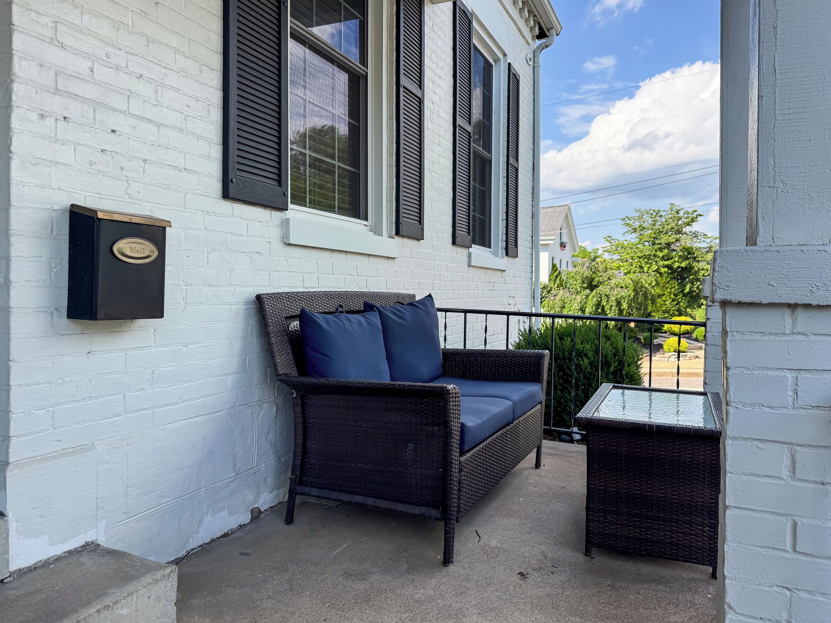 A wicker sofa with cushions, a mailbox, and a small table on a balcony with a white brick wall and a window with shutters.