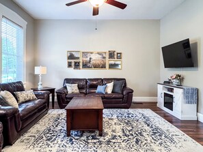 A living room with a brown leather sofa, a wooden coffee table, a patterned rug, a ceiling fan, and a mounted TV. - Mini Aussie | Dog-friendly, Fire pit, Fenced yard (Washington)