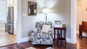 A living room with a patterned armchair, a side table with a magazine, and a refrigerator in the background.