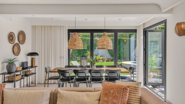 Ein modernes Wohnzimmer mit einer Couch, Esstisch und Stühlen, großen Fenstern mit Blick auf Grünflächen und dekorativen gewebten Lampen.