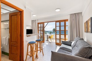 Ein Wohnbereich mit einer Couch, zwei Barhocker, einem Fernseher und einem Balkon mit Meerblick.