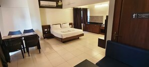 A hotel room with a bed, a desk with chairs, a small kitchenette, and a bathroom. - PRATHAM INN BY DOWNTOWN HOTELS (Manipal)