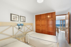 Ein Schlafzimmer mit zwei Betten, einem Holzschrank, gerahmten Bildern an der Wand und Meerblick durch eine offene Tür.