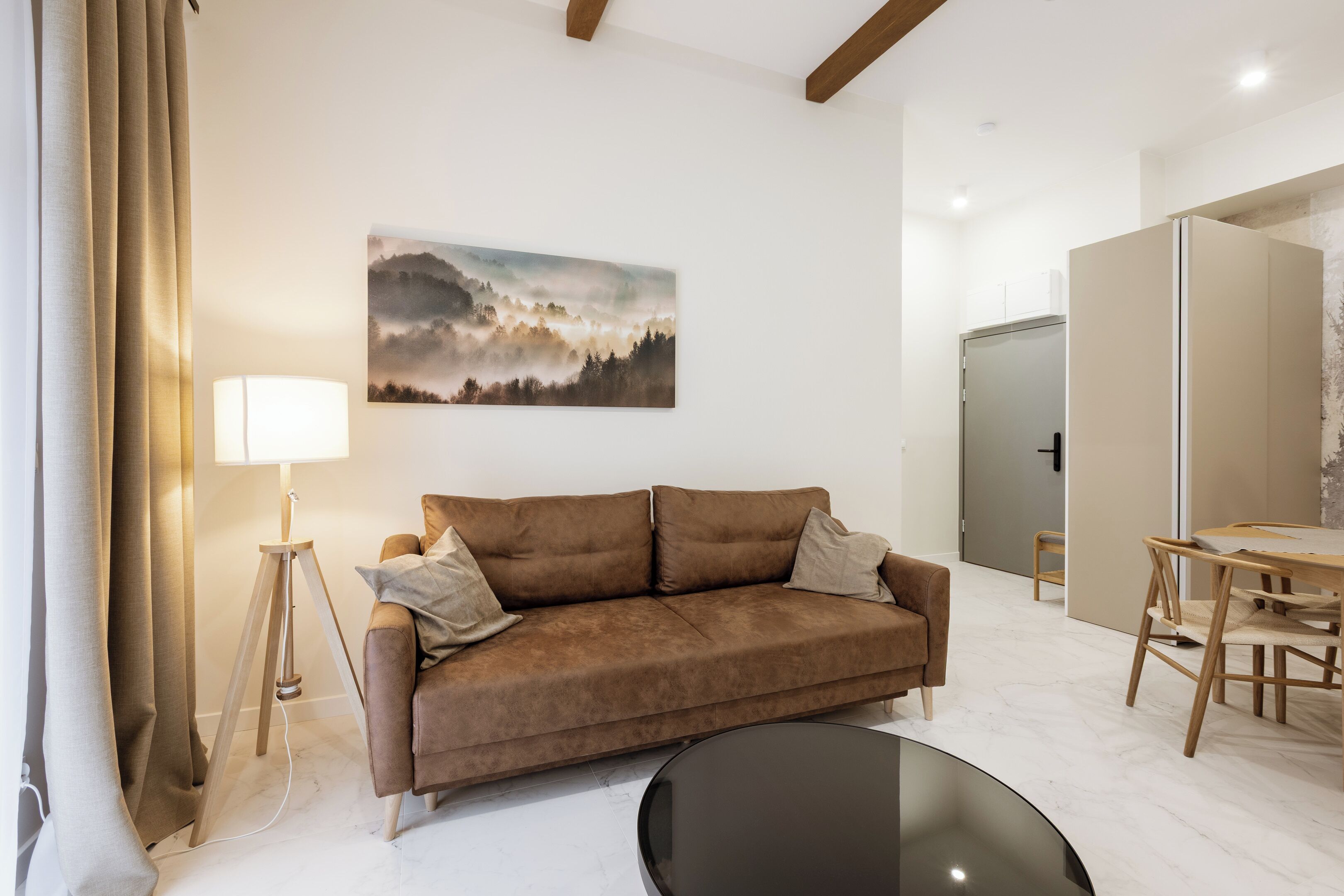 A modern living room with a brown sofa, a round black coffee table, a wooden dining table with chairs, a floor lamp, and a painting of a mountain landscape on the wall.