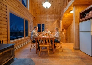 Interior de una cabaña de madera con una mesa de comedor, sillas, un refrigerador y un ventanal grande.