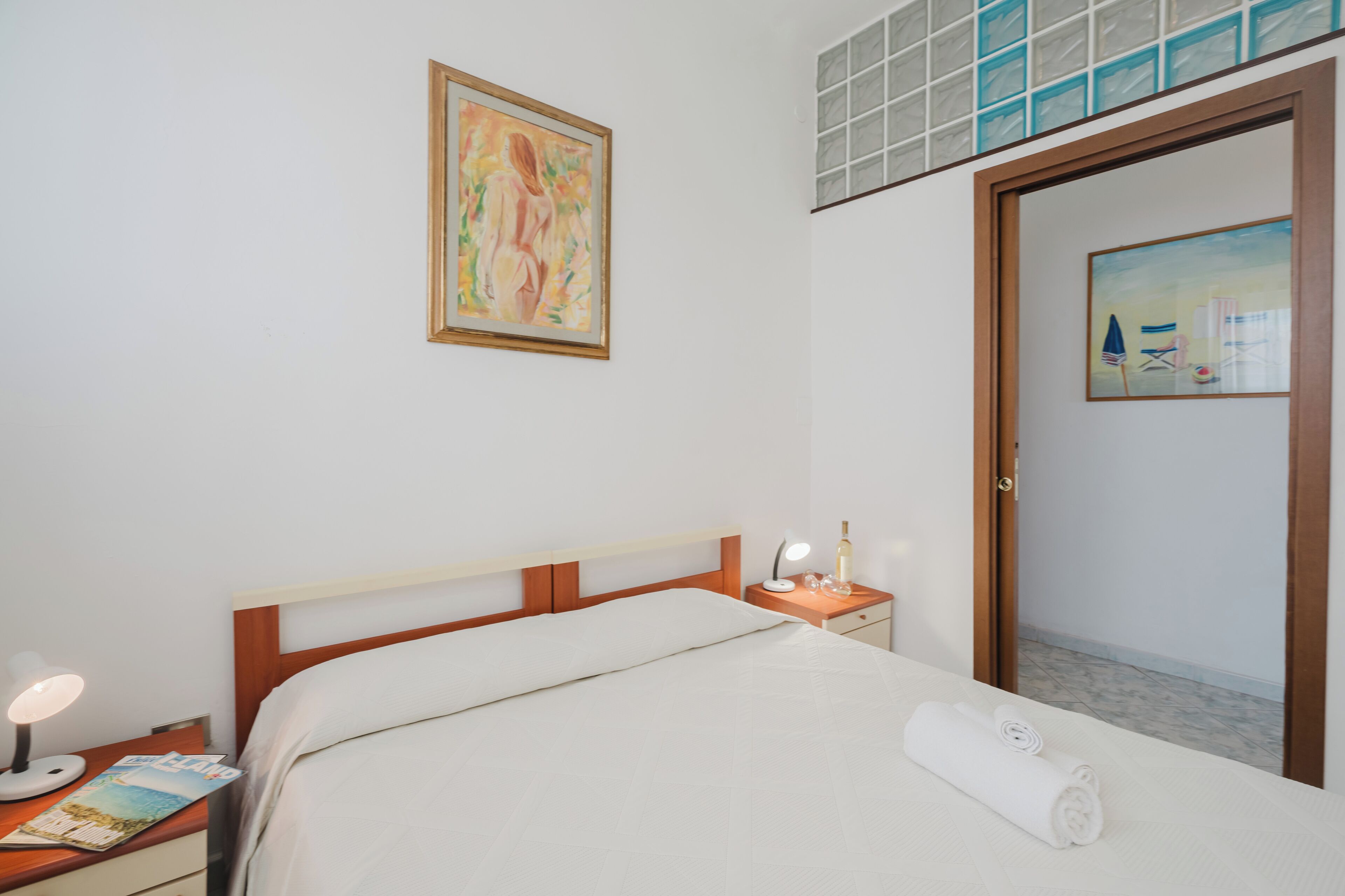 A neatly made bed with a white comforter and a rolled towel, a bedside table with a lamp and a bottle, and a painting on the wall.