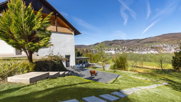 Ein modernes Haus mit Terrasse, Essbereich und Blick auf eine Stadt und Hügel im Hintergrund.