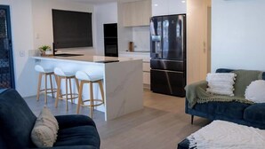 A modern kitchen with a central island, bar stools, a black refrigerator, and a dark blue sofa in a living area. - Cerulean Beachfront - Unit 1 (Sunshine Coast)