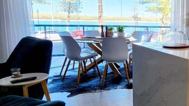A modern dining area with a large window overlooking a scenic view, a round table with chairs, and a small side table with a remote control. - Cerulean Beachfront - Unit 1 (Sunshine Coast)