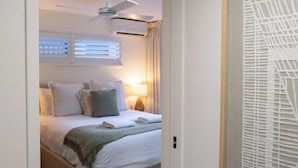 A hotel room with a bed, pillows, a bedside lamp, an air conditioning unit, and a window with blinds. - Cerulean Beachfront - Unit 1 (Sunshine Coast)