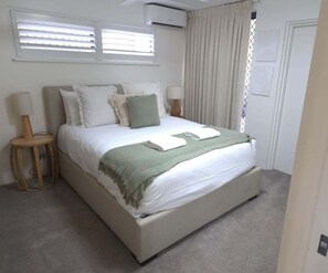 A bedroom with a bed, two bedside tables, a window with blinds, and a door. - Cerulean Beachfront - Unit 1 (Sunshine Coast)