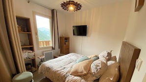 Een slaapkamer met een bed, een televisie, een houten nachtkastje, een raam met gordijnen en een plank met spullen.