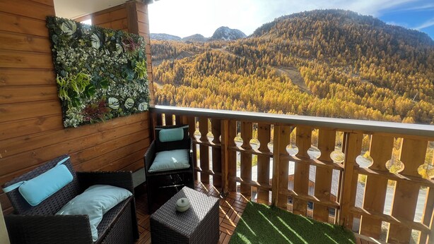 Een balkon met rieten meubilair, kunstgras en uitzicht op een bebost berglandschap.