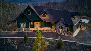Uma casa grande de dois andares, com fachada de pedra e telhado verde, cercada por um jardim bem cuidado e uma paisagem montanhosa.