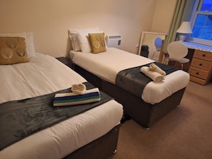 A hotel room with two beds, each with a pillow and a towel. There is a desk with a lamp and a chair, and a window with curtains. - The Longchamp Retreat of Ely (Ely)