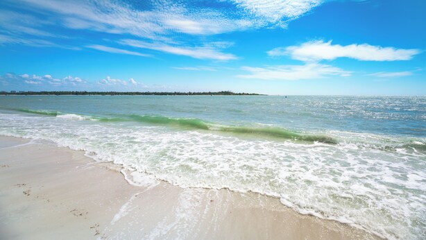 Ein Strand mit Wellen, blauem Himmel und einer fernen Küste.