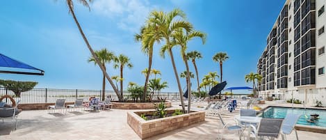 A poolside area with lounge chairs, palm trees, and a building with balconies.