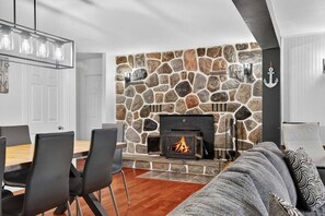 A modern living room with a stone fireplace, a dining area with chairs, and a sofa with patterned cushions. - Bécassines | Private Spa | Billiards | Waterfront (Nominingue)