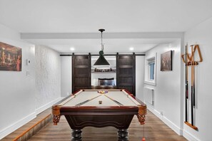 A billiards table in a room with a wooden floor, a hanging light fixture, and a door with a dark wood finish. - Bécassines | Private Spa | Billiards | Waterfront (Nominingue)