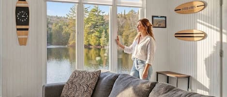 A room with a large window overlooking a lake, a grey sofa, and wooden decor elements.