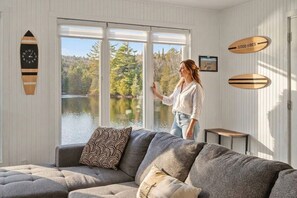 A room with a large window overlooking a lake, a grey sofa, and wooden decor elements. - Bécassines | Private Spa | Billiards | Waterfront (Nominingue)